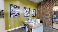 The business center offers a well-appointed workspace that balances functionality with contemporary design. The room features a sleek, stone-topped workstation accommodating multiple users, equipped with LG desktop computers and ample desk space for productivity. Ergonomic wood-backed chairs provide comfortable seating for extended work sessions.

The design aesthetic is a striking combination of bold chartreuse-green walls and an exposed brick accent wall, creating a dynamic visual contrast that energizes the space. Three large black-and-white abstract artworks are displayed prominently above the workstation, adding a sophisticated artistic touch.

The carpeted flooring complements the overall color palette, while bright overhead lighting ensures the workspace remains well-lit throughout the day. The compact yet efficiently organized layout maximizes the available space without feeling cramped. The blend of industrial elements, such as the exposed brick, with modern furnishings and vibrant color choices gives the business center a distinctive character that is both professional and visually engaging.