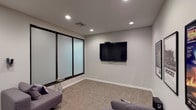 The Media Center is a thoughtfully designed single-level space built for comfortable viewing and relaxed entertainment. The room centers around a large wall-mounted flat-screen television, positioned to provide clear sightlines from multiple seating areas. Several modern gray lounge chairs and compact sofas are arranged throughout the carpeted space, offering flexible and inviting places to sit.

The flooring features a textured, neutral-toned carpet tile that adds warmth underfoot while complementing the light gray walls. Recessed ceiling lighting provides even, ambient illumination throughout the room, creating a relaxed yet functional atmosphere.

A prominent design feature is the large frosted glass partition wall framed in black, which spans a significant portion of one wall. This element adds a sleek, contemporary aesthetic to the space while subtly separating areas within the room.

Adding personality and a cinematic theme, the walls are decorated with a curated collection of framed classic movie posters, including iconic titles such as Star Wars, Rocky, Back to the Future, Jaws, and The Godfather. A subwoofer speaker is also present, suggesting an enhanced audio setup. The overall ambiance is casual, modern, and well-suited for media viewing and entertainment.