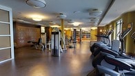 This fitness center offers a spacious, single-floor layout designed to accommodate a full range of workout needs. The space is outfitted with a robust selection of Life Fitness strength-training machines and cable systems, complemented by a row of treadmills and additional cardio equipment positioned along one side. The flooring consists of dark, durable rubber matting suited for heavy equipment and high-traffic use.

Overhead, a grid of circular flush-mount and drum-style light fixtures provides consistent, warm illumination throughout the space, creating a welcoming yet focused environment. Structural columns finished in a warm gold tone are distributed across the floor, adding to the overall aesthetic while delineating workout zones.

A large glass partition wall at the far end of the space opens the visual field, revealing an additional workout or studio area beyond — enhancing the sense of depth and openness. Wall-mounted mirrors further amplify the spatial quality and allow users to monitor their form during exercise.

The warm-toned wall paneling and refined design details elevate the space beyond a standard gym, giving it the feel of an upscale, hotel-caliber fitness facility. Overall, the space strikes a balance between functional utility and polished design.