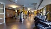 This fitness center offers a spacious, single-floor layout designed to accommodate a full range of workout needs. The space is outfitted with a robust selection of Life Fitness strength-training machines and cable systems, complemented by a row of treadmills and additional cardio equipment positioned along one side. The flooring consists of dark, durable rubber matting suited for heavy equipment and high-traffic use.

Overhead, a grid of circular flush-mount and drum-style light fixtures provides consistent, warm illumination throughout the space, creating a welcoming yet focused environment. Structural columns finished in a warm gold tone are distributed across the floor, adding to the overall aesthetic while delineating workout zones.

A large glass partition wall at the far end of the space opens the visual field, revealing an additional workout or studio area beyond — enhancing the sense of depth and openness. Wall-mounted mirrors further amplify the spatial quality and allow users to monitor their form during exercise.

The warm-toned wall paneling and refined design details elevate the space beyond a standard gym, giving it the feel of an upscale, hotel-caliber fitness facility. Overall, the space strikes a balance between functional utility and polished design.