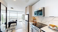 This efficiently designed one-bedroom, one-bathroom unit offers a thoughtful layout that maximizes every square foot across a single floor. The open-concept living and sleeping area features striking exposed concrete ceilings that lend an industrial character, complemented by light wood flooring and a neutral color palette throughout. A wall of floor-to-ceiling windows floods the space with natural light and frames expansive city and skyline views, creating an airy, open atmosphere despite the compact footprint.

The kitchen is sleek and modern, outfitted with stainless steel appliances including a refrigerator, electric range, built-in microwave, and dishwasher. White upper cabinetry is paired with warm wood lower cabinets, and a marble-style backsplash adds an elegant touch to the cooking space.

The generously sized bedroom accommodates a full sleeping area alongside a dedicated workspace, and includes a sliding-door closet for convenient storage. Built-in shelving provides additional organizational flexibility.

The combined bathroom and laundry room features a wood-toned vanity, large-format tile flooring, a walk-in shower with white subway tile, and a stacked in-unit washer and dryer — offering notable practicality within the space. The overall design balances modern finishes with functional, efficient living.