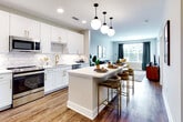 Located in the heart of Charlotte, NC, this single-floor residence at 101 W Morehead St offers a thoughtfully designed living space with a cohesive modern aesthetic throughout. The open-concept kitchen is a standout feature, showcasing white shaker cabinets with gold hardware, a white quartz countertop island, marble-style subway tile backsplash, a gold-finish sink faucet, and globe pendant lighting overhead. Stainless steel appliances, including a French door refrigerator, complete the well-equipped space. Rich hardwood-style flooring flows seamlessly from the kitchen into the living area, which is bathed in natural light from large windows dressed with deep teal drapes. The living room offers a welcoming atmosphere with direct access to an outdoor area. The bedroom features plush carpeting, a ceiling fan, generous window views, and ample closet space, providing a comfortable and restful retreat. The bathroom is finished with classic black-and-white mosaic tile flooring, white cabinetry with gold accents, a large framed mirror, stylish vanity lighting, and a glass-enclosed walk-in shower clad in white subway tile with gold and matte black fixtures — a refined blend of contemporary and timeless design.