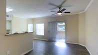 This single-floor space centers around a bright, open living area that combines thoughtful design with functional features. Dark hardwood-style flooring runs throughout, providing a clean contrast to the light-toned walls and white crown molding that frames the room with a polished finish. A ceiling fan with an integrated light fixture is centrally mounted, offering both airflow and ambient lighting to the space.

Natural light is a defining characteristic of this living area, with a large sliding glass door fitted with vertical blinds serving as the primary focal point of the exterior wall. The door provides direct access to an outdoor area, while an adjacent window further enhances the brightness of the room. Together, these openings allow sunlight to pour across the flooring, creating an airy and welcoming atmosphere.

A partial view of a raised countertop or breakfast bar is visible along one side, suggesting a connected kitchen or dining area nearby. White baseboards complete the clean aesthetic throughout. The overall ambiance is bright, modern, and well-suited for comfortable everyday living.
