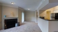 This single-floor home offers a welcoming living area characterized by neutral, warm-toned walls and soft wall-to-wall carpeting that adds comfort underfoot. The focal point of the room is a classic fireplace framed in dark tile with a white mantel, lending a cozy and traditional feel to the space. Crown molding runs along the upper perimeter of the room, adding an elegant architectural detail that enhances the overall finish. A textured ceiling complements the clean white trim and baseboards throughout. A sliding glass door with vertical blinds allows for natural light and likely provides access to an outdoor area. The open layout connects the living area to an adjacent space, visible through a partial wall with a dark countertop, suggesting a pass-through to a kitchen or bar area. A doorway leads further into the home, hinting at additional rooms beyond. The single-floor layout ensures convenient, step-free living throughout. The combination of classic finishes, functional design, and warm tones creates a comfortable and inviting atmosphere well-suited for everyday living.