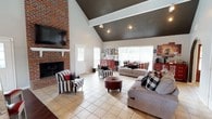 Located at 715 Rice Rd in Ridgeland, Mississippi, this single-story home makes a striking first impression with its dramatic vaulted ceilings and bold architectural character. The expansive living room serves as the centerpiece of the home, showcasing soaring cathedral ceilings with dark-painted upper vaults and exposed white beams that add depth and contrast. A full-height brick fireplace anchors one wall, complemented by a classic white mantle and a wall-mounted television, creating a warm and inviting focal point. Large windows allow natural light to fill the space, while tile flooring throughout the living area adds a clean, durable finish. The kitchen is a standout feature, outfitted with crisp white shaker-style cabinetry, granite countertops, rich dark hardwood flooring, and a full suite of stainless steel appliances including a double oven range, built-in microwave, and refrigerator. A peninsula with seating and pendant lighting provides casual dining options and connects the kitchen seamlessly to the rest of the living space. An arched doorway adds an elegant architectural detail, transitioning smoothly between the main living and kitchen areas. The overall design blends rustic warmth with modern updates, resulting in a cohesive and functional single-floor layout.
