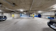 This underground parking facility offers a spacious, single-level layout designed to accommodate a significant number of vehicles, including cars, trucks, and motorcycles. The structure is built with robust concrete construction throughout, with exposed concrete ceilings, walls, and support columns — many of which are marked with high-visibility yellow safety striping to guide drivers and pedestrians safely through the space. Overhead fluorescent strip lighting ensures consistent illumination across the entire facility.

The parking area is divided into distinct zones, each with concrete flooring and clearly delineated driving lanes. A secured gated entry point, fitted with a metal roll-up gate, controls vehicle access to the facility. Overhead, an exposed network of pipes and conduits runs throughout, characteristic of a well-maintained underground structure.

A designated waste management area is located within the facility, equipped with blue recycling bins and a blue dumpster, supporting organized waste disposal. Interior access doors connect to the building above, providing convenient pedestrian entry and exit. Additional utility and storage areas are present within the space. The facility's scale and layout make it well-suited for serving a mid-to-large residential or commercial building, offering practical, secure, and sheltered parking for occupants.