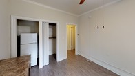 This single-floor unit offers a clean, functional layout with a cohesive aesthetic defined by neutral cream-toned walls, wood-look plank flooring, and classic crown molding throughout. The main living area is generously proportioned, featuring two windows that invite natural light and offer views of surrounding greenery. A ceiling fan with a light fixture adds comfort and practicality to the space. A built-in countertop desk with cabinetry below provides useful storage and workspace, making the room versatile for various uses. Wall-mounted heater units are present in multiple rooms, offering efficient climate control. A secondary room provides additional flexible space, also finished with the same wood-look flooring and crown molding, with its own window and wall heater. A dedicated closet with a bifold door and built-in shelving offers convenient storage, situated alongside a full-size refrigerator. The bathroom is neatly appointed with a toilet, pedestal sink, and a shower/tub combination with a curtain rod, all finished in bright white. Throughout the space, the consistent use of white trim, neutral walls, and warm flooring creates a cohesive and welcoming atmosphere.
