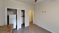 This single-floor unit offers a clean, functional layout with a cohesive aesthetic defined by neutral cream-toned walls, wood-look plank flooring, and classic crown molding throughout. The main living area is generously proportioned, featuring two windows that invite natural light and offer views of surrounding greenery. A ceiling fan with a light fixture adds comfort and practicality to the space. A built-in countertop desk with cabinetry below provides useful storage and workspace, making the room versatile for various uses. Wall-mounted heater units are present in multiple rooms, offering efficient climate control. A secondary room provides additional flexible space, also finished with the same wood-look flooring and crown molding, with its own window and wall heater. A dedicated closet with a bifold door and built-in shelving offers convenient storage, situated alongside a full-size refrigerator. The bathroom is neatly appointed with a toilet, pedestal sink, and a shower/tub combination with a curtain rod, all finished in bright white. Throughout the space, the consistent use of white trim, neutral walls, and warm flooring creates a cohesive and welcoming atmosphere.