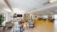 Located at 1513 Versailles Rd in Lexington, Kentucky, this single-floor community space offers a generously sized, open-plan layout finished with light wood-look flooring and a drop panel ceiling with recessed lighting throughout. The main gathering area is divided into a comfortable lounge zone and a dedicated dining area. The lounge features multiple seating arrangements including sofas, armchairs, and a large wooden coffee table, complemented by floor lamps, potted plants, and a wall-mounted flat-screen TV atop a rustic console with open shelving. Large windows along the perimeter flood the space with natural light and provide views of the surrounding greenery. The dining section accommodates multiple tables and chairs, suitable for group gatherings, and is adorned with framed black-and-white equestrian photography — a nod to Lexington's celebrated horse country heritage. A fully equipped kitchen rounds out the space, featuring dark espresso cabinetry with gold hardware, expansive white quartz countertops, stainless steel appliances including a range with a commercial-style hood, wall ovens, and a large refrigerator. The overall ambiance is warm, modern, and functional, making this an ideal venue for community events and activities.