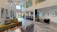 Located at 7599 Eldorado Pkwy in Frisco, TX, this contemporary community clubhouse is designed to impress with its striking double-height ceilings and expansive, open-concept layout. The main lounge area is anchored by large clerestory windows that flood the space with natural light, complemented by wood-look tile flooring in a rich, multi-toned finish that runs throughout. Seating arrangements feature modern mid-century-inspired chairs and sofas in muted teal, gray, and olive tones, accented with patterned throw pillows for a curated, welcoming feel. A dramatic globe chandelier serves as a focal point overhead, adding an artistic touch to the open gathering area. The space transitions seamlessly into a bar-style counter with bar stools and pendant lighting, creating an ideal spot for socializing. A built-in booth with upholstered seating and a round table provides a more intimate setting nearby. Framed abstract artwork adorns the walls, while wall-mounted televisions offer entertainment throughout. A billiards table is tucked into one corner, adding a recreational element to the space. Black-framed glass partition walls and doors contribute to the sleek, industrial-modern aesthetic, allowing visual connectivity between zones while defining distinct areas within this thoughtfully designed community gathering space.