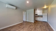 This single-floor unit offers a practical and clean layout designed for comfortable everyday living. The living area is open and inviting, finished with warm wood-look laminate flooring that extends throughout the space, complemented by neutral gray walls and crisp white trim. A wall-mounted mini-split air conditioning unit provides efficient climate control. The kitchen is positioned adjacent to the living area and features white cabinetry, dark granite-style countertops, a white refrigerator, a range with an overhead vent hood, and a single-basin sink — offering ample storage and workspace for home cooking. The bedroom is a well-proportioned, private space with a large window fitted with blinds that brings in natural light while maintaining privacy. The full bathroom is finished with large-format beige ceramic tile surrounding a soaking tub and shower combination, a white vanity with under-cabinet storage, a medicine cabinet mirror, and tile flooring. A front entry door with an adjacent window provides additional natural light to the main living space. The unit's cohesive finishes and functional design make it a comfortable and efficient living environment throughout.