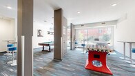 This contemporary amenity space is designed as a fully equipped game room and social lounge, offering a dynamic environment for recreation and entertainment. The open-plan layout is anchored by a classic pool table positioned beneath a stylish pendant light fixture, surrounded by ample floor space for gameplay. A Super Chexx bubble hockey table and a foosball table add to the variety of entertainment options available throughout the room.

A dedicated shuffleboard area is set apart from the main gaming zone, accompanied by wall-mounted flat-screen televisions and bar-height stools, creating an ideal spot for spectators. Modern bar stools with teal upholstery and sleek chrome bases are distributed throughout the space, complementing the patterned carpet flooring that features bold teal accents woven into a grey base.

Floor-to-ceiling windows along one wall flood the interior with natural light and provide open views of the surrounding exterior. The neutral white and taupe wall palette, punctuated by contrasting structural columns, gives the space a clean, modern aesthetic. A glass-panel entryway connects the lounge to adjacent corridors, ensuring seamless flow. The overall ambiance is energetic yet polished, making this a well-appointed communal gathering space suited for active social interaction.