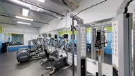 This fitness facility is designed to support a comprehensive workout experience within a single-level layout. The space is outfitted with a solid selection of cardio equipment, including Precor elliptical machines and stationary bikes, each equipped with individual monitor screens for tracking performance metrics. A multi-station cable machine is prominently positioned on the floor, offering a range of strength training options for users of varying fitness levels.

Full-length mirrors line the walls, visually expanding the space and allowing users to monitor their form during exercise. The walls feature a clean white base accented by vibrant green and blue horizontal color bands, lending the space an energetic and motivating atmosphere. Overhead, exposed ductwork and industrial-style ventilation run along the ceiling, contributing to a utilitarian aesthetic that keeps the focus on function.

The flooring consists of durable, dark rubber matting, well-suited for heavy equipment and high-traffic use. Fluorescent lighting ensures the space is brightly and evenly lit throughout. The overall layout is organized and practical, providing users with clear pathways between equipment and a comfortable environment for fitness activities.
