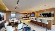 This sophisticated single-floor amenity lounge is designed to serve as a premier social hub, combining multiple entertainment and gathering functions within one thoughtfully appointed space. The open-plan layout is anchored by a spacious social lounge area furnished with a large navy sectional sofa, cream swivel accent chairs, and a bold navy, gold, and white area rug that adds a striking visual focal point. Floor-to-ceiling windows flood this area with natural light and offer views of the outdoor pool terrace beyond.

A full billiards table occupies its own dedicated zone, complemented by wall-mounted flat-screen televisions and warm wood-paneled accent walls that lend the space a refined, club-like atmosphere. Pendant lighting throughout enhances the upscale feel.

The fully equipped kitchenette features sleek wood cabinetry, white quartz countertops, stainless steel appliances, and a large mirrored backsplash that visually expands the space. An adjacent bar-height island with white upholstered counter stools provides an ideal setting for casual dining and socializing.

A semi-private media room enclosed by glass partition walls offers a more intimate retreat, furnished with a plush navy sofa and a dedicated flat-screen television. Near the entrance, a stylish dining area with a round glass table and upholstered chairs sits beside a dramatically textured dark accent wall. Large-format light tile flooring ties the entire space together seamlessly.