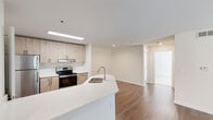 Located on Wesley Place in Lawrenceville, Georgia, this single-floor unit offers a clean, contemporary design throughout. The open-concept living area is finished with warm hardwood-style flooring and neutral white walls, creating a bright and inviting atmosphere. A sliding glass door fills the space with natural light while providing direct access to an outdoor area overlooking lush, mature trees.

The kitchen is a functional centerpiece of the unit, featuring light wood cabinetry, a classic subway tile backsplash, stainless steel appliances including an electric range and refrigerator, and a large peninsula with a white quartz-style countertop and an integrated sink. This layout supports both everyday cooking and casual entertaining.

The bedroom is carpeted for comfort and includes a window with blinds that allows ample natural light, along with access to a closet and adjoining spaces. The bathroom is finished in a clean, all-white palette with a full tub-and-shower combination, white tile surround, and chrome fixtures.

The exterior, as seen in the point-cloud scan, reveals a multi-story building set amid a generously vegetated landscape, providing a peaceful, nature-adjacent environment for residents.