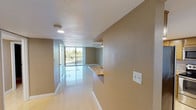 This single-floor space offers a clean, open layout designed to maximize both functionality and natural light. The interior is finished with large, light-toned tile flooring that runs throughout, reflecting ambient light and lending a bright, airy feel to the space. Textured walls in warm neutral tones provide a subtle contrast to the crisp white baseboards and ceilings, which feature a classic popcorn finish with recessed lighting.

A defining architectural highlight is the expansive floor-to-ceiling window wall at the far end of the space, allowing abundant natural light to pour in and offering open views of the surrounding greenery and exterior environment. This glazed wall creates a strong visual connection to the outdoors, enhancing the sense of openness within.

A peninsula or pass-through counter with a stone or marble surface is visible along one side, suggesting a functional separation between the kitchen and living areas while maintaining the open feel of the floor plan. A ceiling fan is also present, providing both practical ventilation and a casual finishing touch to the living area. The overall ambiance is clean, neutral, and well-lit — well-suited for a variety of interior configurations and uses.