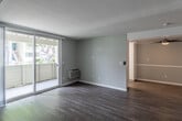 Located at 10280 La Hacienda Ave in Fountain Valley, California, this single-story residence offers a well-organized layout finished with wood-style flooring throughout. The open living area is bright and airy, anchored by a wall-unit air conditioner and a sliding glass door that leads to a private balcony with views of surrounding greenery, allowing natural light to flow freely into the space. A secondary room, also fitted with a ceiling fan and its own sliding glass door to an outdoor area, provides flexible living options.

The galley-style kitchen features white cabinetry, light countertops, stainless steel appliances including an electric range and dishwasher, and a dual-basin sink beneath a window with horizontal blinds, creating a clean and functional workspace. The primary bedroom includes a ceiling fan and connects directly to a private bathroom with a double-sink vanity and a glass-enclosed shower. Additional bedrooms are equipped with ceiling fans and windows dressed in blinds. A second bathroom offers a tub-and-shower combination with a curved rod and a bright vanity. Textured ceilings are present throughout the main living areas, complementing the neutral gray palette that lends the space a clean, contemporary feel.