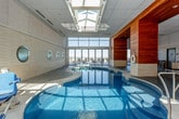 This thoughtfully designed indoor aquatic facility combines striking architectural elements with practical amenities to create a refined swimming environment. At the heart of the space is a generously sized freeform indoor pool with a smooth curved shape, complemented by an adjacent hot tub. The pool deck is finished in polished concrete, providing a clean, durable surface that surrounds the water on all sides.

Natural light plays a central role in the space's ambiance. A large skylight runs along the ceiling above the pool, flooding the interior with daylight, while a dramatic floor-to-ceiling glass wall and sliding glass doors open views to an exterior terrace and the surrounding landscape beyond. Along one side, a series of distinctive circular porthole windows are set into a textured stone-clad wall, adding a nautical design element and allowing additional light to filter through.

Warm, horizontal wood paneling anchors a key section of the space, providing visual contrast against the lighter stone and concrete surfaces. A lounge and seating area is situated adjacent to the pool, furnished with tables and chairs for relaxation. An accessibility lift is positioned at the pool's edge, ensuring inclusive access to the water. The overall design balances contemporary aesthetics with a welcoming, resort-like atmosphere ideal for both recreation and leisure.