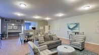 This single-story space showcases an open-concept layout that seamlessly connects the living, dining, and kitchen areas, creating a cohesive and functional environment. The living area is furnished with a plush gray sofa and coordinating patterned accent chairs arranged around a round coffee table, providing a comfortable and well-organized seating arrangement. A large abstract canvas painting in soft blue and green tones serves as a focal point on one wall, complementing the neutral color palette of the space.

The flooring throughout is a warm hardwood-style material that adds continuity across the open layout. Recessed flush-mount ceiling fixtures provide consistent overhead lighting throughout the space. The dining area is positioned adjacent to the living room and features a round table with upholstered chairs, partially visible through an architectural opening that leads toward the kitchen.

The kitchen area, visible in the background, features white cabinetry and dark countertops. Walls are painted in complementary shades of beige and soft blue-gray, contributing to the calm, neutral ambiance. A high-top bar table with stools near one wall adds a casual seating option. Overall, the space presents a clean, modern aesthetic with thoughtful furnishings and a cohesive design sensibility.