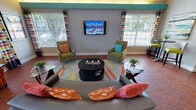 This single-story communal space combines functional design with a lively, energetic aesthetic. Upon entry, a welcoming foyer greets visitors with terracotta tile flooring, soaring vaulted ceilings fitted with ceiling fans, and a decorative console table accented by colorful artwork. French doors and transom windows flood the entrance with natural light.

The reception area features two sleek, textured dark-paneled workstations beneath a dropped ceiling with accent lighting and track spotlights, creating a polished and professional environment. A hallway connects the reception zone to additional areas within the space.

The lounge area is generously proportioned, with high vaulted ceilings and a bold teal accent ledge running along the upper walls with integrated uplighting. A wall-mounted flat-screen television serves as a focal point, flanked by large windows dressed in vibrant polka-dot curtains with blinds. An arched clerestory window adds an architectural detail while maximizing daylight. Seating includes patterned accent chairs, striped armchairs, and a gray sofa arranged around a circular black coffee table atop a colorful area rug. A high-top bistro table and bar stools provide an additional casual seating option, rounding out a dynamic and inviting communal environment.