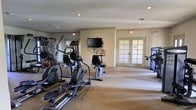 This single-floor fitness center offers a practical and functional workout environment designed to accommodate a range of exercise needs. The space is finished with neutral beige walls and carpeted flooring, creating a clean and understated aesthetic throughout. Recessed ceiling lights provide consistent, even illumination across the room, supplemented by natural light that filters in through a set of elegant French doors along the far wall, which also serve as the primary entry and exit point. The cardio equipment area is prominently featured and includes multiple elliptical trainers and a stationary bike arranged in a row, allowing for comfortable spacing between machines. A wall-mounted flat-screen television is positioned centrally, providing entertainment for those using the cardio equipment. Additional strength or resistance training equipment is visible along the perimeter of the room. Emergency exit signage is clearly posted, reflecting attention to safety standards. The open layout of the room allows for ease of movement between equipment, and the overall design balances functionality with a welcoming, low-key ambiance suited for everyday fitness use.