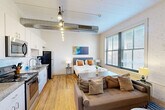 Located at 915 Locust St in the heart of downtown St. Louis, this efficiently designed studio unit offers a thoughtfully arranged open-plan living space on a single floor. Warm hardwood floors run throughout the main area, complementing the bright, neutral walls and elevated ceilings that lend a sense of openness. Exposed industrial ductwork adds a loft-style character, while a striking multi-bulb track light fixture anchors the kitchen zone. Large windows flood the space with natural light and frame views of the surrounding urban streetscape.

The kitchen is well-appointed with white shaker cabinetry, granite countertops, a subway tile backsplash, and a full suite of stainless steel appliances including a refrigerator, range, microwave, and dishwasher. The open layout seamlessly connects the kitchen to the combined living and sleeping area, which comfortably accommodates a full-sized bed, a sofa, and a dining table with chairs.

A hallway with a louvered door provides access to the bathroom, which features grey tile flooring, a modern vanity with granite countertop, a framed mirror, and clean, contemporary fixtures. The overall ambiance is modern and functional, blending industrial architectural elements with polished, comfortable finishes.