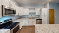 This single-floor unit at County Road D E in Maplewood, Minnesota, offers a well-designed open layout with a clean, contemporary aesthetic throughout. The kitchen serves as a central highlight, featuring crisp white shaker cabinetry, granite countertops, a stainless steel appliance suite including a refrigerator, range, microwave, and dishwasher, and a kitchen island with a teal accent base. Dark hardwood-style flooring runs through the kitchen and living areas, adding warmth and contrast to the light blue-gray walls.

The spacious living area extends toward a large sliding glass door that opens to a private balcony overlooking lush, mature trees, filling the space with natural light. Two bedrooms are finished with plush carpeting, windows with wooded views, and closet access, offering a quiet and comfortable retreat.

The unit includes two bathrooms, both featuring granite-topped white vanities, hardwood-style flooring, and modern chrome fixtures. One bathroom includes a soaking tub and shower combination, while the other offers a walk-in shower. A dedicated laundry area is also present within the unit. Overall, the space presents a bright, functional, and well-appointed living environment.