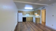 This single-floor space is designed with functionality and practicality in mind, offering a straightforward layout suited for comfortable everyday living. The kitchen serves as a central feature, equipped with warm-toned wood cabinetry that lines the walls in an L-shaped configuration, providing ample storage and counter space. The kitchen includes essential appliances such as a white refrigerator, a white electric range, and a black dishwasher, alongside a sink positioned beneath upper cabinetry. A flush-mounted ceiling light fixture illuminates the kitchen area, ensuring a bright and well-lit workspace. The space is grounded by rich, dark hardwood-style flooring that runs throughout, adding warmth and visual continuity to the interior. The walls are finished in a clean, neutral white tone, contributing to a bright and open atmosphere. Ceiling details include a subtle overhead beam accent, adding a mild architectural element to the room. A visible HVAC vent is integrated into the ceiling, ensuring climate comfort throughout. A doorway leads to an adjoining area, suggesting additional living space beyond the kitchen. Overall, this unit presents a clean, no-frills environment with a focus on livability and practicality.