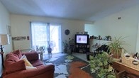 This single-floor living space centers around a comfortable, casual living room that blends functionality with a homey atmosphere. The room features a textured ceiling and neutral-toned walls that provide a clean backdrop for the eclectic décor. A large sliding glass door along one wall floods the space with natural light and offers a view of an outdoor patio area, where additional greenery can be seen. Vertical blinds frame the door, allowing for flexible light control. The living area is anchored by a bold, large-scale floral area rug in earth tones of rust, green, and gray, adding a strong visual focal point to the carpeted floor. Seating includes a deep red upholstered sofa, contributing a warm accent to the room. An entertainment center houses a television and media components, complemented by decorative items displayed on nearby shelves. The space is notably enriched by an abundance of indoor plants, positioned throughout the room, adding life and a natural, organic quality to the interior. A wall clock and various small personal accents contribute to the lived-in, welcoming character of the space.