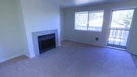 This single-floor residential space presents a clean and functional layout designed for comfortable everyday living. The living area serves as the heart of the home, featuring wall-to-wall carpeting that adds warmth and a sense of coziness underfoot. A standout feature of the room is the fireplace, framed by a classic tile surround, which anchors the space and provides a natural focal point. The neutral-toned walls and light-colored carpet create a bright, open atmosphere that feels welcoming and versatile, easily adaptable to a variety of interior styles. A large window fitted with horizontal blinds allows ample natural light to fill the room while offering views of the surrounding neighborhood, contributing to an airy and connected feel. The room's generous proportions provide plenty of space for furniture arrangement, making it well-suited for both relaxing and entertaining. Wall outlets are conveniently positioned, and the clean baseboard trim throughout adds a polished finishing touch. Overall, the space offers a straightforward, move-in-ready environment with practical features and a calm, neutral aesthetic that serves as a solid foundation for personalization.