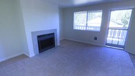 This single-floor residential space presents a clean and functional layout designed for comfortable everyday living. The living area serves as the heart of the home, featuring wall-to-wall carpeting that adds warmth and a sense of coziness underfoot. A standout feature of the room is the fireplace, framed by a classic tile surround, which anchors the space and provides a natural focal point. The neutral-toned walls and light-colored carpet create a bright, open atmosphere that feels welcoming and versatile, easily adaptable to a variety of interior styles. A large window fitted with horizontal blinds allows ample natural light to fill the room while offering views of the surrounding neighborhood, contributing to an airy and connected feel. The room's generous proportions provide plenty of space for furniture arrangement, making it well-suited for both relaxing and entertaining. Wall outlets are conveniently positioned, and the clean baseboard trim throughout adds a polished finishing touch. Overall, the space offers a straightforward, move-in-ready environment with practical features and a calm, neutral aesthetic that serves as a solid foundation for personalization.