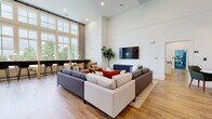 Located at 3506 Buster Lane in Indian Land, SC, this community club room at Madison Place is a thoughtfully designed, single-floor amenity space that blends modern aesthetics with practical functionality. The interior is defined by soaring ceilings and crisp white walls that create a bright, airy atmosphere, complemented throughout by warm-toned hardwood flooring.

The main lounge area serves as the centerpiece of the space, featuring a generous sectional seating arrangement, wall-mounted television, and an adjacent sitting nook with accent chairs — ideal for relaxed gatherings. Tall, multi-pane windows flood the room with abundant natural light and offer views of the surrounding greenery.

A built-in kitchenette is outfitted with white shaker cabinetry, a subway tile backsplash, a central island with bar seating, a stainless steel sink, and a dishwasher — well-suited for entertaining and events. Decorative open shelving adds both style and utility to the kitchen area.

A dedicated co-working station runs along a floor-to-ceiling window wall, featuring a long wooden countertop with individual desk lamps and bar-height seating, providing an inspiring, light-filled workspace. Additional casual seating vignettes are arranged throughout, accented by pendant lighting and curated wall art. A corridor leads to further amenities, including what appears to be a fitness area.