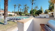 This inviting outdoor community space showcases a stunning resort-style pool area defined by its Spanish Colonial architectural influence. The main swimming pool is generously sized and surrounded by a broad, tiled pool deck lined with rows of lounge chairs, offering ample space for relaxation under open blue skies. Vibrant green shade umbrellas are positioned throughout, providing welcome relief from the sun.

Towering date palms and mature trees frame the entire area, creating a lush, tropical atmosphere that enhances the sense of a private retreat. The architecture features characteristic white stucco walls with warm golden trim, decorative arched colonnades, and ornate ironwork gates — all hallmarks of the Spanish Colonial Revival style. A gated entry clearly marked as the Main Swimming Pool ensures a controlled, secure environment for residents.

A dedicated barbecue station is integrated into the space, complete with a posted rules board and surrounded by tropical plantings including large-leafed banana plants and ornamental shrubs. Decorative lamp posts add charm and provide lighting throughout the grounds. The surrounding multi-story residential buildings, with their terra cotta-tiled roofs and balconies, complement the cohesive architectural aesthetic. The overall ambiance is serene, well-maintained, and evocative of a warm-climate resort destination.