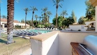 This inviting outdoor community space showcases a stunning resort-style pool area defined by its Spanish Colonial architectural influence. The main swimming pool is generously sized and surrounded by a broad, tiled pool deck lined with rows of lounge chairs, offering ample space for relaxation under open blue skies. Vibrant green shade umbrellas are positioned throughout, providing welcome relief from the sun.

Towering date palms and mature trees frame the entire area, creating a lush, tropical atmosphere that enhances the sense of a private retreat. The architecture features characteristic white stucco walls with warm golden trim, decorative arched colonnades, and ornate ironwork gates — all hallmarks of the Spanish Colonial Revival style. A gated entry clearly marked as the Main Swimming Pool ensures a controlled, secure environment for residents.

A dedicated barbecue station is integrated into the space, complete with a posted rules board and surrounded by tropical plantings including large-leafed banana plants and ornamental shrubs. Decorative lamp posts add charm and provide lighting throughout the grounds. The surrounding multi-story residential buildings, with their terra cotta-tiled roofs and balconies, complement the cohesive architectural aesthetic. The overall ambiance is serene, well-maintained, and evocative of a warm-climate resort destination.