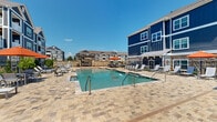 Located at 3262 S Railroad St in Phenix City, Alabama, this outdoor amenity space serves as a resort-style retreat designed for relaxation and recreation. The centerpiece is a generously sized swimming pool with crystal-clear water, accessible via multiple entry points including a pool lift for accessibility. The pool is surrounded by an expansive paver deck finished in warm-toned stone, providing ample room for lounging and socializing. Numerous sling-style chaise lounge chairs are arranged throughout the deck, complemented by shaded seating areas furnished with tables and market umbrellas in a bold orange hue. A charming poolside cabana structure, clad in stone and topped with a pitched shingle roof, offers a shaded gathering spot complete with a pergola accent. The space is enclosed by sleek black metal fencing, with thoughtfully placed landscaping adding greenery and color along the perimeter. The backdrop features a striking multi-story residential building with bold navy blue board-and-batten siding, stone base accents, and white-trimmed balconies. Under the open Alabama sky, this outdoor space delivers an inviting, well-appointed atmosphere ideal for warm-weather enjoyment.