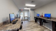 This single-floor business center is designed to provide a functional and comfortable workspace for its users. The room features two long, granite-topped counters lining opposite walls, each equipped with multiple desktop computers, keyboards, and high-backed bar-style chairs, offering ample workstation space for several users simultaneously. The flooring throughout is a light-toned wood-look tile, lending the space a clean and modern aesthetic. The walls are painted in a calming two-tone scheme of soft blue-gray on the upper half and a lighter neutral below, contributing to a focused yet welcoming ambiance. Natural light filters in through multiple windows fitted with white blinds, as well as through a glass-paneled exterior door, keeping the space bright and airy. A casual seating nook near the exterior door features a round natural-edge table paired with metal hairpin-leg stools, offering an informal area for conversation or relaxed work. Overhead fluorescent lighting provides consistent illumination throughout. A second interior doorway with a pass-through window connects to an adjacent area, suggesting the business center is part of a larger facility. Overall, the space is thoughtfully laid out to balance productivity and comfort.