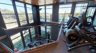 This well-appointed two-story fitness center combines modern design with comprehensive workout amenities. The ground floor features durable rubber flooring throughout its strength training area, equipped with a variety of cable machines, free weights, and functional training equipment. Ceiling fans and recessed lighting keep the space comfortable and well-lit. A dedicated spin studio with hardwood floors and full-length mirror walls houses a row of indoor cycling bikes, offering an energizing environment for cycling workouts. A separate flexibility and stretching area with plush carpet, wall-mounted TVs, and stability ball storage provides a quieter space for functional fitness. An open staircase with wood-and-metal railing connects to the upper level, where large steel-framed windows wrap around the perimeter, flooding the space with natural light and offering sweeping views of the surrounding landscape. The upper floor features a warm wood-paneled ceiling, adding a refined touch to the cardio zone, which is lined with treadmills, ellipticals, and rowing machines. Throughout both levels, the dark charcoal wall palette, industrial-style exposed ductwork, and floor-to-ceiling glazing create a sleek, contemporary aesthetic that is both functional and visually striking.