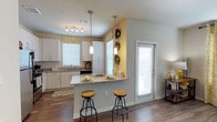 This single-floor space showcases a well-designed open-concept kitchen and living area that maximizes both functionality and style. The kitchen is appointed with crisp white cabinetry featuring sleek hardware, granite countertops, and a peninsula breakfast bar that seats two, making it an ideal casual dining and entertaining hub. Two pendant lights hang above the bar, complementing a track lighting fixture overhead to create layered, warm illumination throughout the space.

Rich hardwood-style flooring runs continuously through the kitchen and into the adjoining living area, grounding the space with a warm, cohesive aesthetic. Multiple windows dressed with patterned roman shades allow generous natural light to fill the interior, contributing to an open and airy atmosphere. A full-light door panel further enhances brightness and provides convenient access to the exterior.

Neutral wall tones in soft gray and warm yellow create a welcoming contrast, while decorative wall accents add a personalized touch. The layout flows naturally between the kitchen and living spaces, making the home feel connected and practical for everyday living.