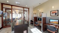 This single-floor business center offers a thoughtfully designed environment catering to both focused work and group collaboration. The space is divided into several functional zones, each serving a distinct purpose while maintaining a cohesive aesthetic defined by warm wood tones, neutral wall colors, and patterned carpeting throughout.

The business center area features a row of semi-private computer workstations, each equipped with desktop monitors and separated by panel dividers. Pendant lighting illuminates each station, while framed artwork adorns the walls above, adding a polished touch to the workspace.

Adjacent to the workstations, a dedicated conference room accommodates a sizable group around a dark rectangular table with mixed seating. A wall-mounted flat-screen television and large windows with roller shades provide both functionality and natural light. A built-in kitchenette with wood cabinetry, granite countertops, under-counter refrigerator, microwave, and tile backsplash supports the meeting area.

A separate lounge nook features two cream leather armchairs arranged around a glass side table, accompanied by a wall-mounted television and sun-filled windows. Glass-paneled doors with wood framing connect the zones, maintaining an open, airy flow throughout the space.