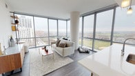This contemporary high-rise unit offers a thoughtfully designed open-concept layout spread across a single floor, combining modern aesthetics with functional living. The defining feature of the living area is the dramatic floor-to-ceiling corner curtain wall windows, which flood the space with natural light and frame expansive panoramic views of the city skyline, green parkland, and a distant waterfront. Wide-plank hardwood-style flooring runs throughout, complementing the clean, neutral palette of the interiors.

The living and dining area flows seamlessly, with the corner glazing creating an airy, light-filled ambiance. The kitchen is sleek and well-appointed, showcasing dark wood cabinetry, a large white waterfall-edge island with bar seating, stainless steel appliances, and pendant lighting — all set against a backdrop of more floor-to-ceiling windows.

The primary bathroom is a refined retreat, featuring a large-format tiled walk-in shower with a frameless glass door, a dark wood vanity with white countertop, and a backlit mirror. A second full bathroom offers a soaking tub with tiled surround, a matching dark vanity, and warm hardwood-style flooring. Together, the spaces present a cohesive, upscale residential environment with strong architectural character and outstanding views.