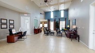 This single-floor communal space makes a strong first impression with its dramatic vaulted, beamed ceilings that rise to a considerable height, creating an open and airy atmosphere throughout. The interior is defined by a bold color palette, combining soft grey walls with a striking teal accent wall that adds depth and visual interest to the space.

Large windows dressed with dark curtain panels allow generous amounts of natural light to filter in, brightening the expansive tile flooring that runs throughout the main area. The lounge zone is thoughtfully arranged with a leather sofa, upholstered armchairs, and an ottoman grouped around a coffee table, providing a comfortable and functional seating area. A decorative ceiling fan complements the open overhead space.

Adjacent areas visible from the main lounge include a dining space and what appears to be a secondary room accessible through glass-paneled doors, suggesting a versatile, multi-use layout. A wooden sideboard near the entrance adds a warm, welcoming touch. Overall, the space conveys an inviting, well-lit, and generously proportioned environment suited for communal gathering and relaxation.