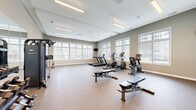Located at 4811 Wildroot Ln in Fredericksburg, Virginia, this fitness center offers a thoughtfully designed, single-floor facility built to support a wide range of workout routines. The space is organized into distinct functional zones, each catering to different training needs.

The main exercise floor is generously proportioned, finished with durable speckled rubber flooring ideal for high-traffic athletic use. Large, multi-pane windows line the walls, flooding the space with abundant natural light and offering views of the surrounding outdoor greenery. Overhead, suspended linear lighting fixtures provide consistent, bright illumination throughout.

This primary area houses a comprehensive selection of cardio equipment, including treadmills, elliptical machines, and stationary bikes, as well as adjustable workout benches and a multi-station cable strength machine. A wall-mounted flat-screen television adds convenience for longer training sessions.

An adjoining functional training room features light hardwood-style flooring and soaring ceilings, creating an open and airy atmosphere. A wall-mounted storage rack is equipped with stability balls, medicine balls, kettlebells, and TRX suspension trainers, making it ideal for group fitness or functional workouts. Pendant lighting and a large window enhance the room's welcoming feel.

Additional support areas, including a transition hallway with wood flooring and safety equipment, complete the facility's practical layout.
