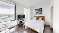 Located at 10 New Street in Boston's East Boston neighborhood, this efficiently designed one-bedroom, one-bathroom unit offers a well-planned layout across a single floor. The space features a clean, contemporary aesthetic throughout, with crisp white walls, dark hardwood-style flooring, and an abundance of natural light.

The bedroom is the standout space, occupying the majority of the unit's footprint. Large floor-to-ceiling windows frame sweeping harbor views, flooding the room with natural light and offering a striking urban waterfront backdrop. The bedroom comfortably accommodates a full sleeping area alongside a living and dining arrangement, making it highly functional.

The kitchen is outfitted with modern grey cabinetry, a stainless steel countertop, a built-in oven, and an over-the-range microwave. Its efficient design maximizes every inch of available space. The bathroom maintains the same modern sensibility, featuring large-format floor tiles, a floating vanity with an integrated sink, and clean white walls accented by contemporary fixtures.

Overall, this unit delivers a sleek, minimalist living experience with high-quality finishes, impressive waterfront views, and a thoughtfully organized layout that makes excellent use of the available space.