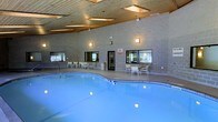 This indoor aquatic facility offers a generously sized swimming pool as its centerpiece, with clear blue water illuminated by underwater lighting that creates a bright, inviting atmosphere. The pool features a curved design and is surrounded by a wide pool deck, providing ample space for movement and relaxation.

The enclosure is constructed with durable concrete masonry block walls, lending the space a solid, institutional character. Above, a warm wood-paneled ceiling with recessed skylights allows natural light to filter in, softening the overall ambiance and complementing the wall-mounted lighting fixtures that keep the space evenly lit throughout.

Seating areas are arranged along the perimeter, with tables and chairs positioned near large windows that offer views to the outdoors, creating a pleasant connection to the surrounding environment. A central entry door provides convenient access to and from the facility.

The single-level layout ensures easy accessibility throughout the space, while the combination of natural and artificial lighting maintains a comfortable, welcoming environment year-round. Whether used for recreational swimming or lap training, this indoor pool facility is designed to be both functional and enjoyable for its users.