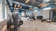 Located on Dynamic Boulevard in Wesley Chapel, FL, this contemporary fitness center offers a spacious, single-floor layout thoughtfully designed to accommodate a full range of workout needs. The main exercise area is defined by a striking teal coffered ceiling accented with diagonal linear LED lighting, creating an energetic and modern atmosphere. Patterned carpet flooring runs throughout the primary training zone, which is outfitted with an extensive collection of Hoist strength machines, free-weight stations including multiple dumbbell racks, adjustable benches, and a cable machine system. A dedicated cardio row lines one side of the space, featuring a full lineup of TRUE treadmills, stair climbers, and elliptical machines positioned beneath a bank of windows that invite ample natural light. Multiple wall-mounted flat-screen TVs are positioned throughout for entertainment during workouts. A separate studio room, enclosed by full-height glass partition doors, offers hardwood flooring, patterned accent walls, ceiling fans, and an array of functional training equipment including kettlebells, medicine balls, resistance bands, and battle ropes. This studio space is ideal for group fitness or individual functional training sessions. Together, the zones create a comprehensive, well-lit, and thoughtfully appointed fitness facility.