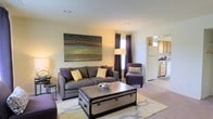 This two-floor model home offers a thoughtfully designed layout with an open-concept main living area that flows seamlessly into the kitchen. The living room features neutral-toned walls and carpeted flooring, creating a warm and inviting atmosphere. A large statement artwork anchors the main wall, complementing the space's earthy color palette of grays, browns, and muted yellows. Natural light filters in through a window dressed with floor-length dark curtains, adding depth and contrast to the bright, clean interior. The living room accommodates a generously sized sofa, an accent chair, and a distinctive trunk-style coffee table that serves as a focal point. Recessed lighting in the ceiling supplements the natural light throughout the day. The adjacent kitchen is visible from the living area and features light wood cabinetry, white appliances, and a functional layout suited for everyday use. The open sightlines between the living and kitchen areas enhance the sense of space, making the home feel connected and practical. The two-floor design further expands the overall living potential of this well-presented model home.