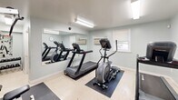 Located at 1314 Wharton Dr in Newark, Delaware, this fitness center offers a well-organized, single-room workout facility designed to accommodate a range of exercise needs. The space features light gray walls, a neutral tile floor, and bright fluorescent lighting that creates a clean and practical atmosphere throughout.

The cardio section is outfitted with treadmills and an elliptical trainer, positioned along one wall and complemented by a large wall-mounted mirror that visually expands the room and allows users to monitor their form. A wall clock is conveniently placed within the mirror area for easy timekeeping during workouts.

On the opposite side of the room, a multi-function cable strength training machine and a weight bench provide options for resistance and strength training. A recumbent bike offers an additional low-impact cardio option. The equipment is arranged on protective rubber floor mats, keeping the space organized and safe.

Additional amenities include a wall-mounted flat-screen television, a water cooler, and a waste bin for user convenience. Windows along the exterior wall allow natural light to enter, while a wall-mounted air conditioning unit ensures climate control. The room is equipped with a smoke detector and an illuminated exit sign, reflecting attention to safety standards.