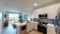 This thoughtfully designed single-floor residence offers a seamless blend of modern style and functional living. The open-concept kitchen and living area serves as the heart of the space, featuring sleek white cabinetry, a peninsula with a stainless steel sink and built-in dishwasher, and a full suite of stainless steel appliances including a French door refrigerator, electric range, and over-the-range microwave. A gray tile backsplash and white quartz countertops complete the kitchen's contemporary aesthetic, while a linear pendant light anchors the space above the peninsula. Light wood-tone flooring flows throughout, unifying the living and kitchen areas. The living room is bright and welcoming, with a large sliding glass door that opens to an outdoor area and fills the room with natural light. The bedroom is a calm retreat, highlighted by a large window overlooking a landscaped outdoor space, a walk-in closet, and light, neutral walls. The bathroom is clean and modern, featuring a walk-in glass shower, a single-sink vanity with white cabinetry, and a wood-framed mirror. Throughout the residence, a cohesive neutral palette and contemporary finishes create a polished, comfortable environment.
