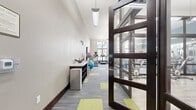 This fitness center offers a thoughtfully designed, multi-zone workout facility spread across a single floor. The space is entered through a striking dark-framed glass door system, immediately revealing a generous, open layout with elevated ceilings that contribute to an airy, energizing atmosphere.

The main exercise floor features patterned carpet tiles in grey and yellow-green, providing a vibrant and durable surface beneath a comprehensive array of cardio equipment, including treadmills and elliptical machines, as well as cable machines, a full dumbbell rack, and various strength training stations. Large multi-pane windows along one wall flood the space with abundant natural light and offer outdoor views, while ceiling fans and suspended linear lighting ensure comfort and visibility throughout.

A dedicated spin studio, enclosed by floor-to-ceiling dark-framed glass partitions, provides a focused cycling environment. This room features light wood-look plank flooring, a wall of mirrors, a mounted flat-screen display, and a ceiling fan, creating an immersive group cycling experience. Additional functional training areas include a stretching and accessories zone stocked with stability balls, medicine balls, and a vertical knee raise station. The overall design balances an open, motivating aesthetic with clearly defined workout zones, making it a comprehensive and well-appointed fitness facility.