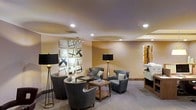 This single-floor space presents a thoughtfully designed lounge area that balances comfort with contemporary style. The room features warm, neutral-toned walls complemented by light hardwood flooring, contributing to an inviting and cohesive aesthetic. Recessed ceiling lighting is distributed evenly throughout, providing consistent illumination, while sleek floor lamps with black shades and gold stands add a layer of ambient warmth and visual interest.

The seating arrangement centers around a collection of plush gray armchairs positioned around a wooden side table, creating an intimate conversation area. Stacked books on the table add a casual, intellectual touch to the setting. Two striking abstract art pieces adorn the walls — one a geometric black-and-white multi-panel composition, the other a vertical piece featuring warm gold and earth tones — lending the space a curated, gallery-like quality.

A subtle tray ceiling detail adds architectural dimension overhead. To one side, a piece of office equipment suggests a functional component to the space. Overall, the room exudes a refined yet approachable atmosphere, well-suited for relaxed gathering or quiet use.